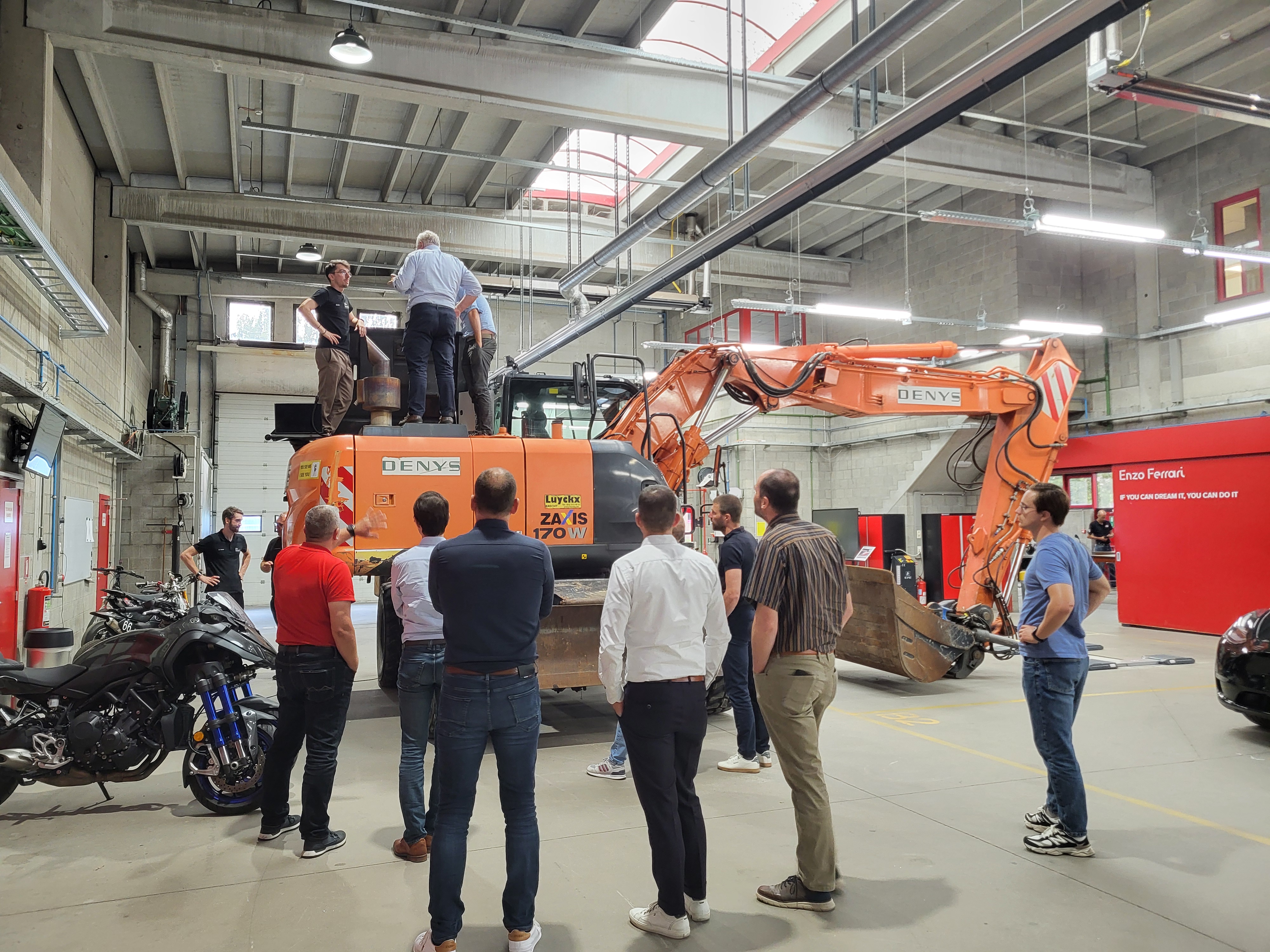 Deelnemers bekijken een oranje graafmachine in het labo tijdens een ReMeWa-projectbijeenkomst.